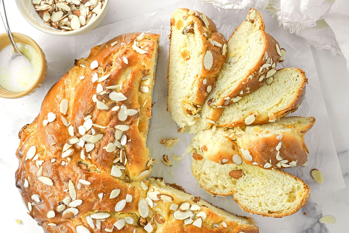 A sliced braided bread loaf topped with slivered almonds sits on parchment paper. Nearby are a small bowl of sliced almonds and a bowl with a spoon containing a white glaze.