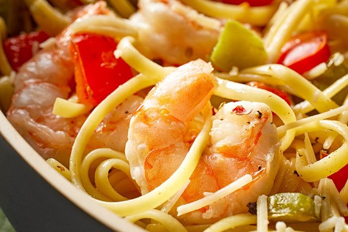 Close-up of cooked shrimp mixed with linguine pasta, diced red and green bell peppers, and sprinkled with herbs in a bowl.