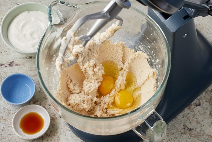Overhead shot of a stand mixer bowl containing creamed ingredients with two raw eggs, paddle attachment in place, surrounded by small bowls of vanilla extract, sour cream, and a light-colored ingredient;