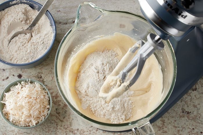 Overhead shot of a stand mixer bowl containing batter, with flour being added and the paddle attachment in place, accompanied by a blue bowl of flour with a spoon and a small bowl of shredded coconut on a light-colored countertop;