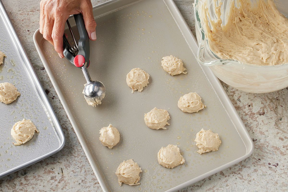 Overhead shot of a hand using a cookie scoop to place balls of cookie dough onto a greased baking sheet, with a bowl of dough and another baking sheet nearby on a countertop;