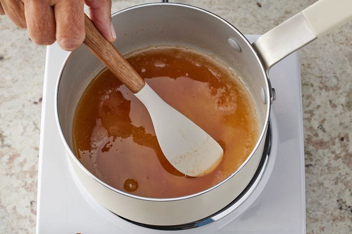 Close shot of a hand stirring a pot of golden-brown liquid, possibly caramel or broth, with a white spatula on a stovetop;