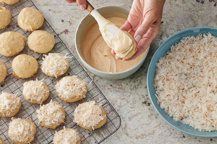 Overhead shot of a person dipping a cookie into icing over a bowl, with iced cookies on a cooling rack and a plate of shredded coconut nearby for coating;