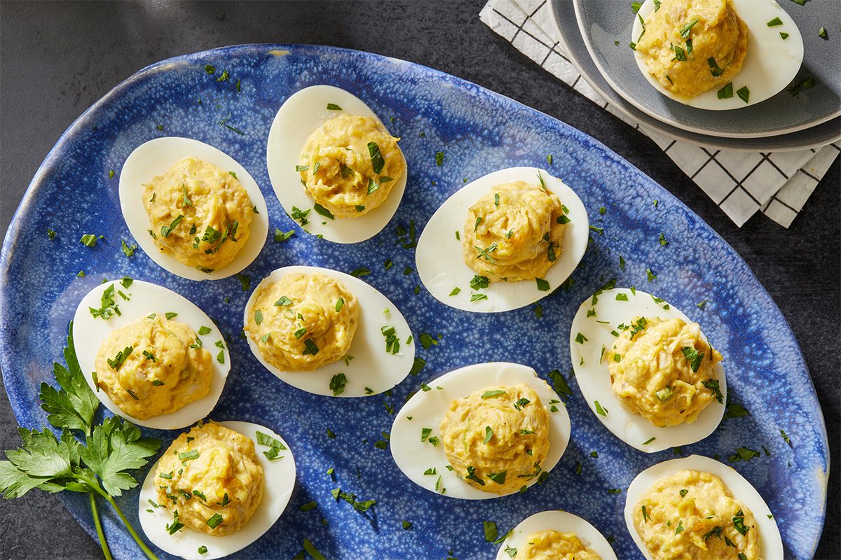 A blue platter with deviled eggs garnished with chopped parsley, and a sprig of parsley beside the platter. A few deviled eggs are served on a small gray plate in the background.