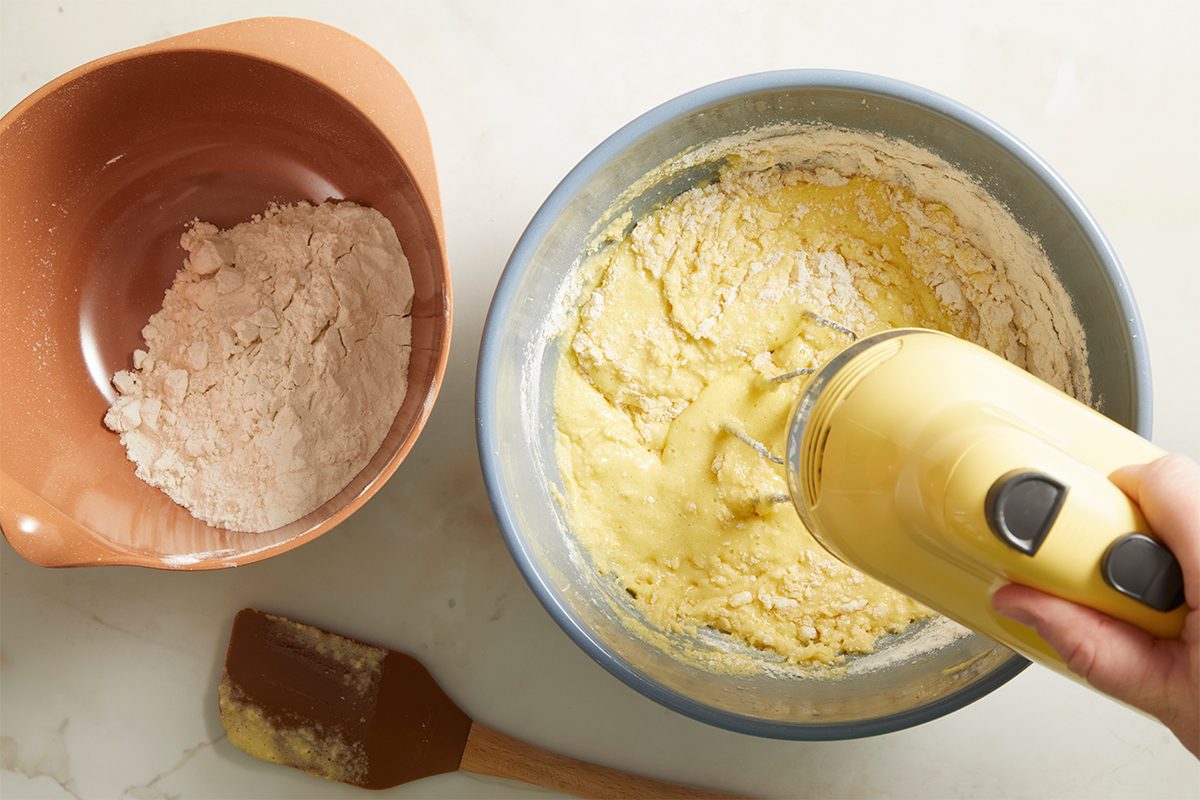 A hand uses a yellow electric mixer to blend batter in a blue bowl, with a brown bowl containing flour and a spatula nearby on a light countertop.
