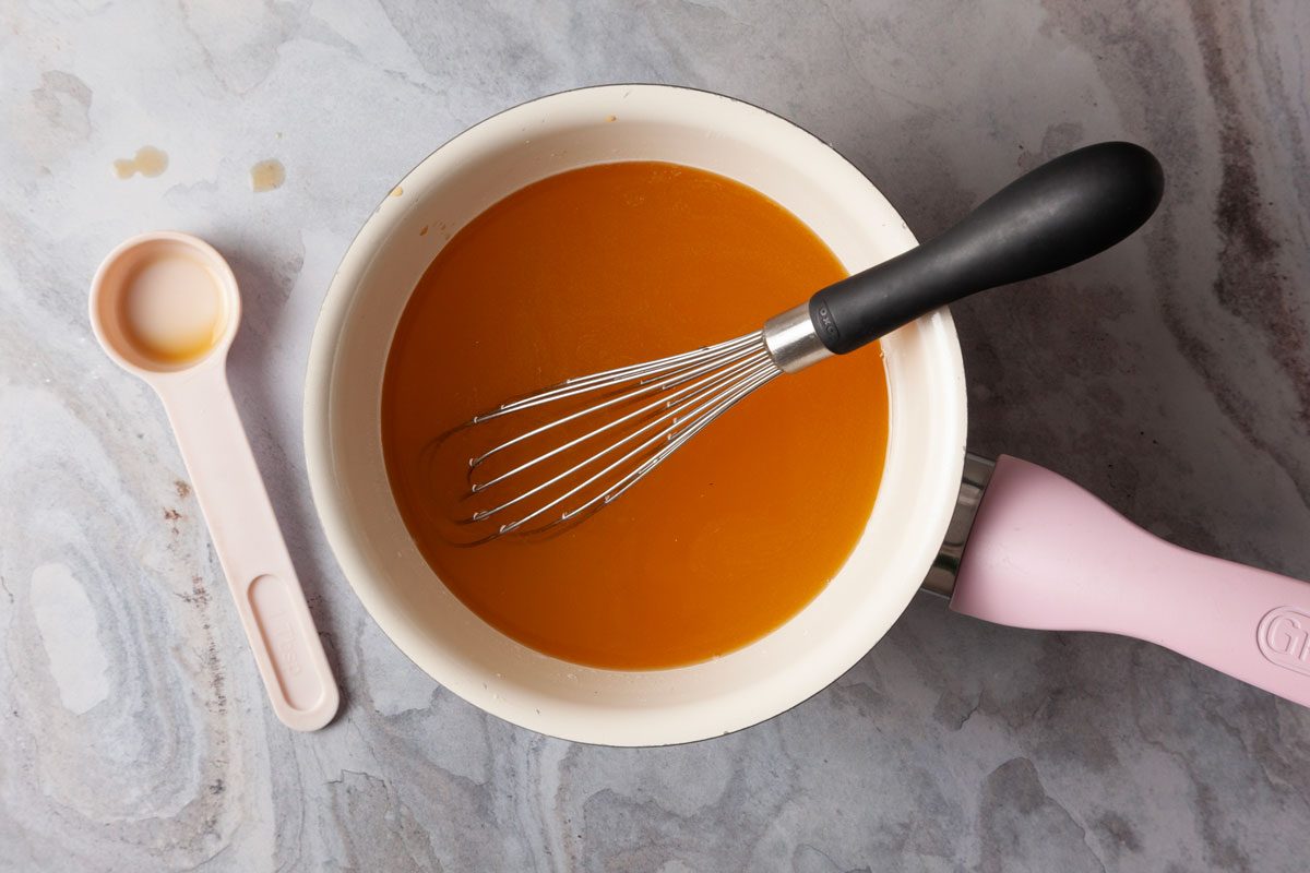 Overhead shot of a saucepan filled with orange-colored liquid and a whisk, placed on a marble countertop beside a pink measuring spoon.