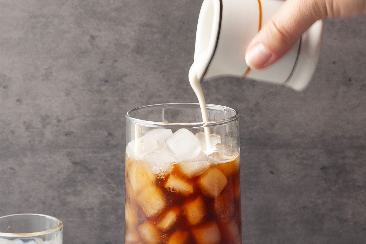 Close shot of a hand pouring cream from a small white pitcher into a tall glass of iced coffee filled with ice cubes, set against a gray background.