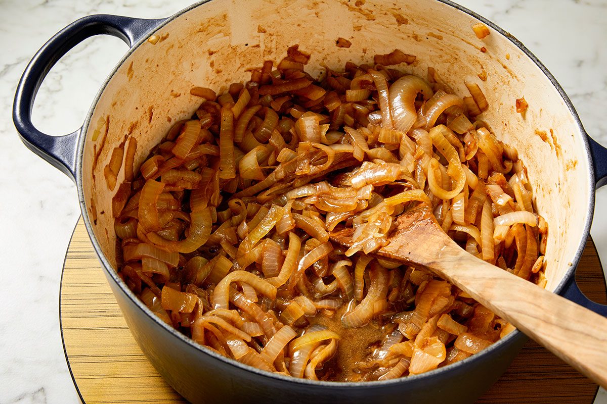 Closeup shot of a pot filled with caramelized onions being stirred with a wooden spoon, placed on a wooden trivet on a light-colored countertop.