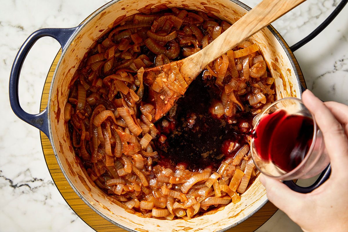 Overhead shot of a hand pouring red wine into a pot of caramelized onions while stirring with a wooden spoon on a marble countertop.