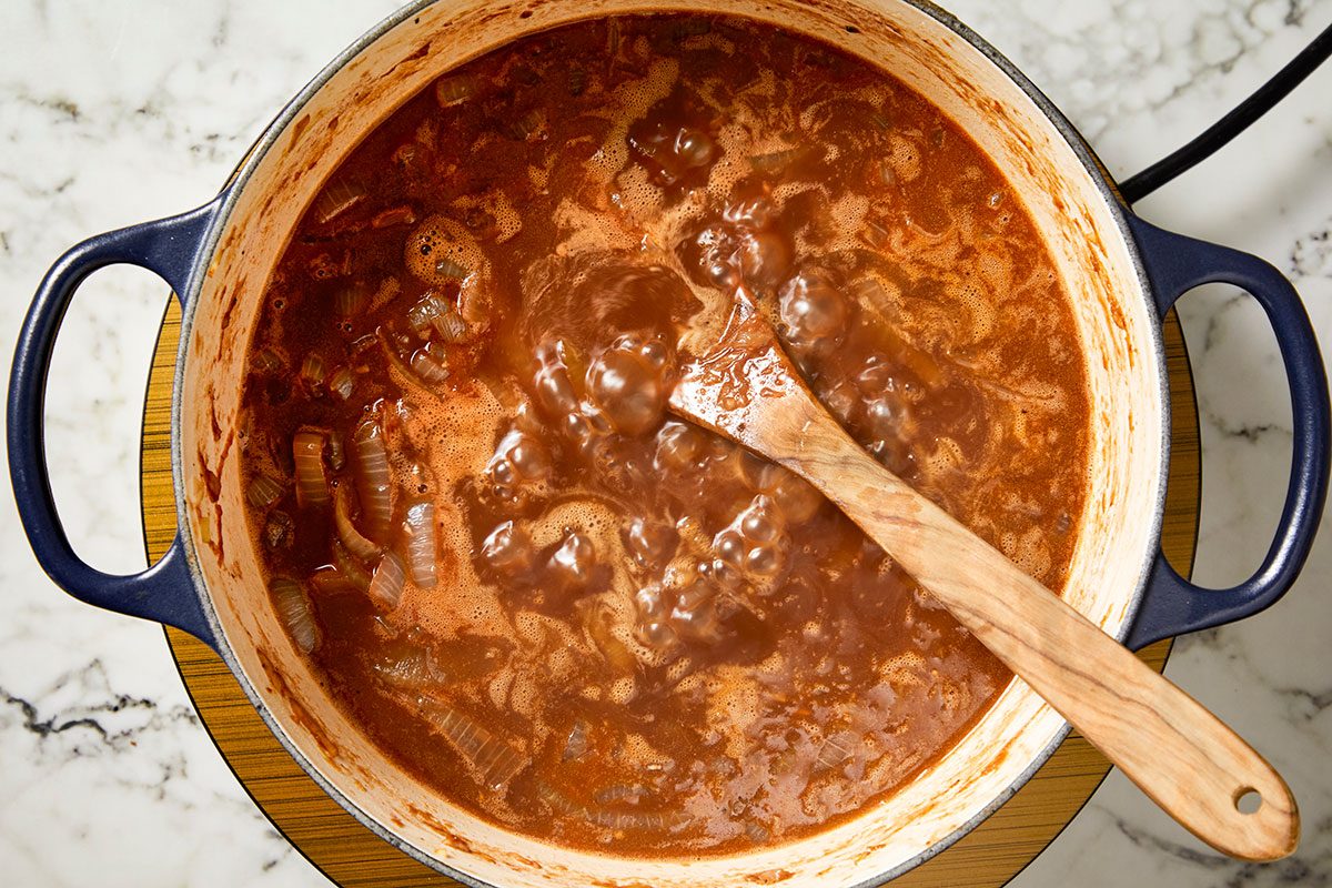 Overhead shot of a blue Dutch oven filled with bubbling red tomato sauce and onions on a marble countertop, with a wooden spoon resting inside the pot;
