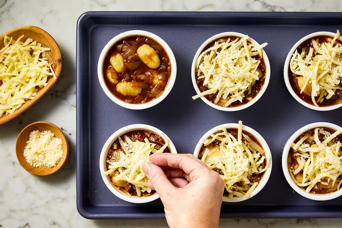 Overhead shot of a hand sprinkling shredded cheese over bowls of soup with gnocchi and vegetables arranged in a baking tray, with two small wooden bowls of grated cheese nearby on a marble countertop;