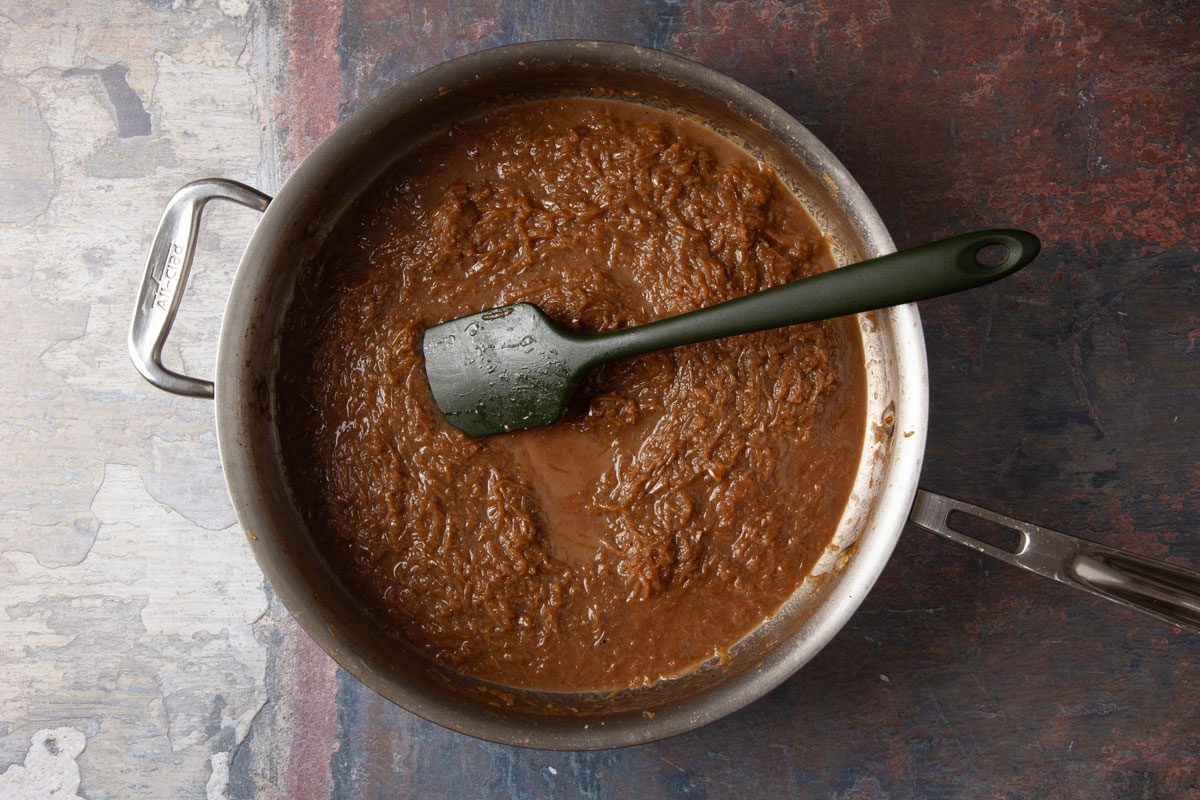 Overhead shot of a metal pan filled with thick brown sauce on a textured surface, with a green spatula resting inside, partially coated in the sauce.