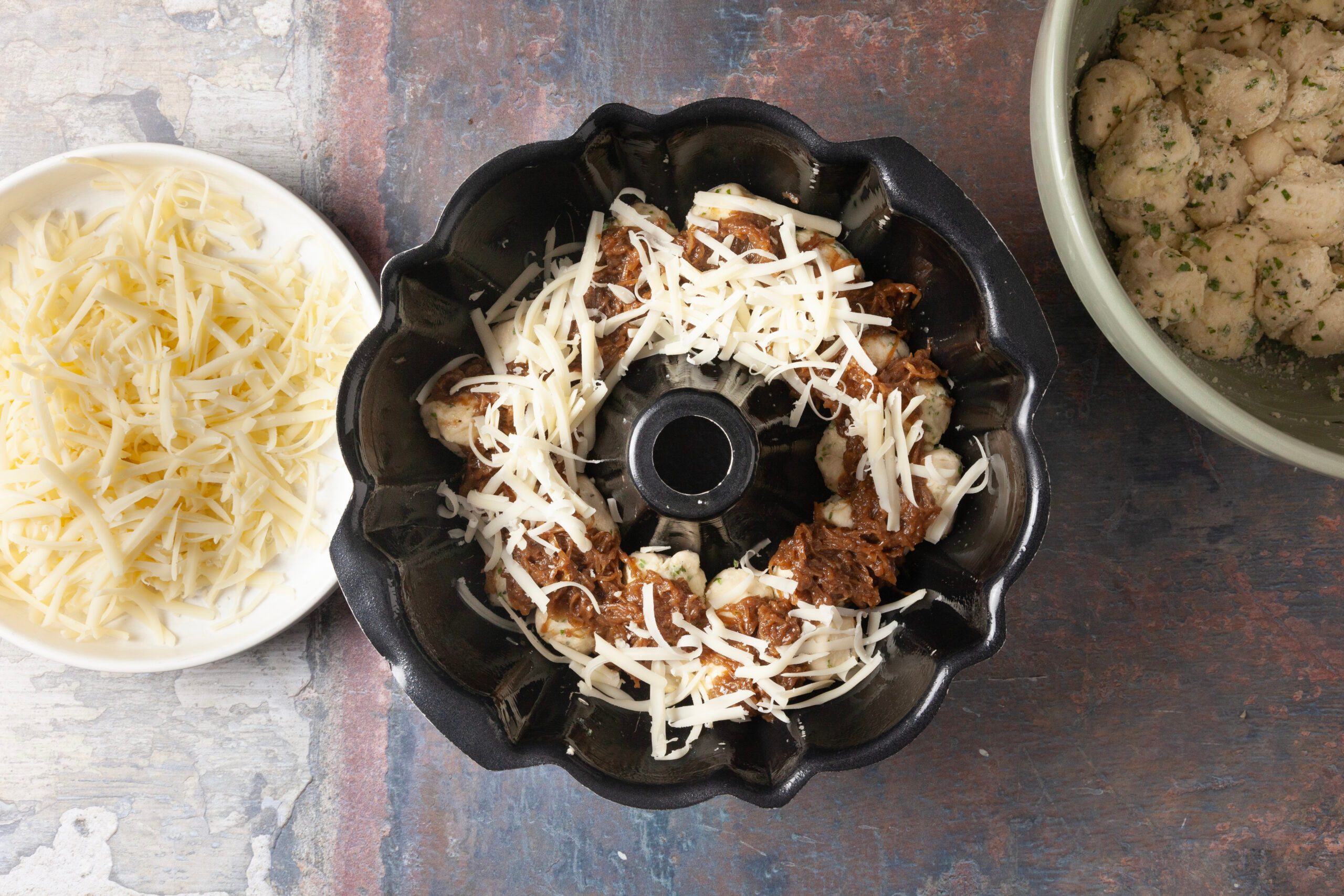 Overhead shot of a bundt pan filled with unbaked monkey bread dough pieces, some coated in seasoning and topped with shredded cheese; To the side, a bowl of shredded cheese and a bowl of herbed dough pieces are visible.