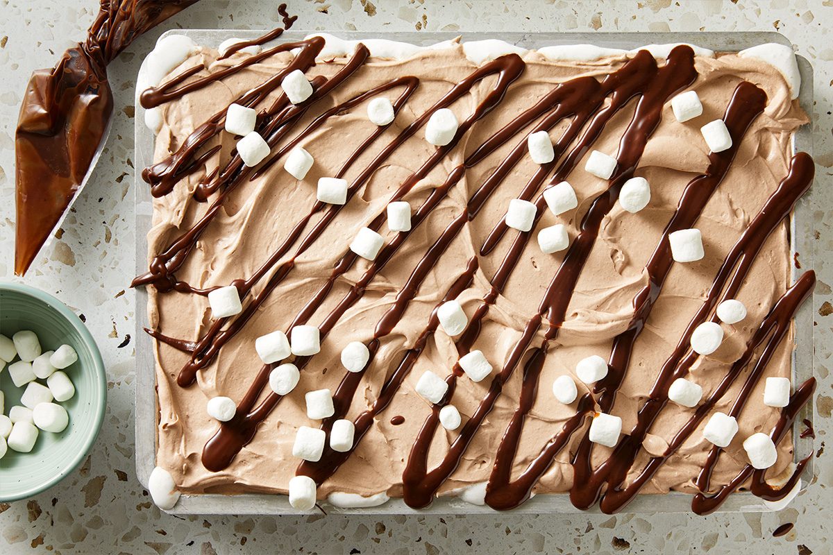 Overhead shot of Hot Chocolate Poke Cake topped with whipped frosting, drizzled chocolate sauce, and mini marshmallows, viewed from above.
