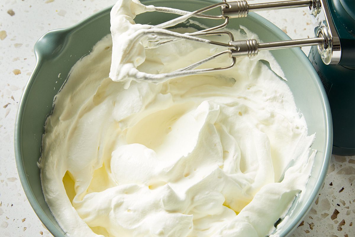 Fluffy whipped cream being prepared in a mixing bowl with electric beaters on a speckled countertop.