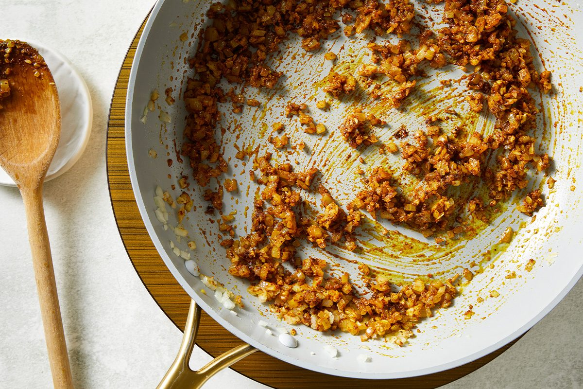 Overhead shot of a sauté pan filled with browned, finely chopped onions and spices, placed on a wooden trivet, with a wooden spoon resting on a white plate nearby;