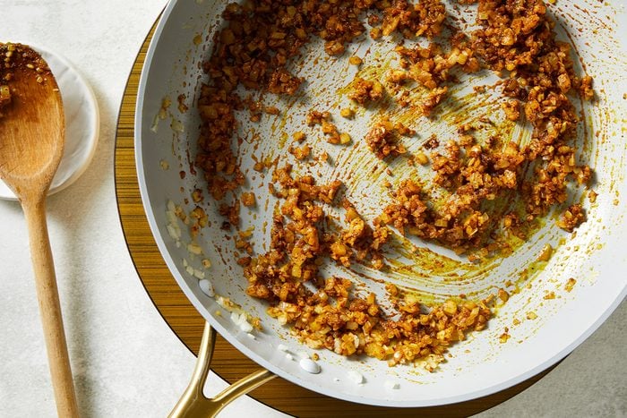 Overhead shot of a sauté pan filled with browned, finely chopped onions and spices, placed on a wooden trivet, with a wooden spoon resting on a white plate nearby;