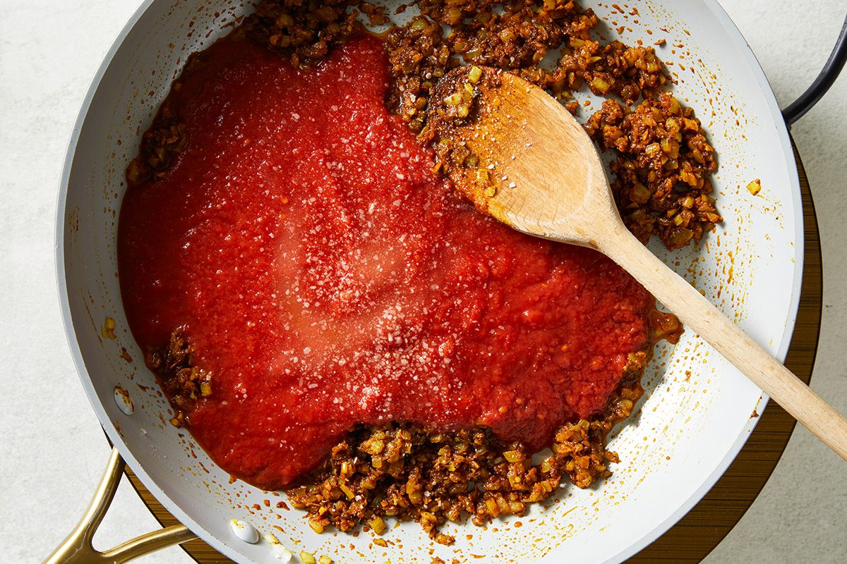 Overhead shot of a skillet containing sautéed vegetables and ground meat, with tomato sauce poured over one side. A wooden spoon rests in the pan, ready to mix everything together;