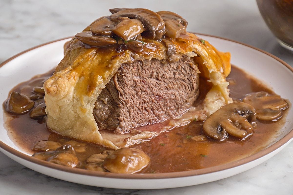 Close shot of a plate with individual Beef Wellingtons cut open, revealing medium beef inside golden pastry, served with sautéed mushrooms and mushroom sauce;