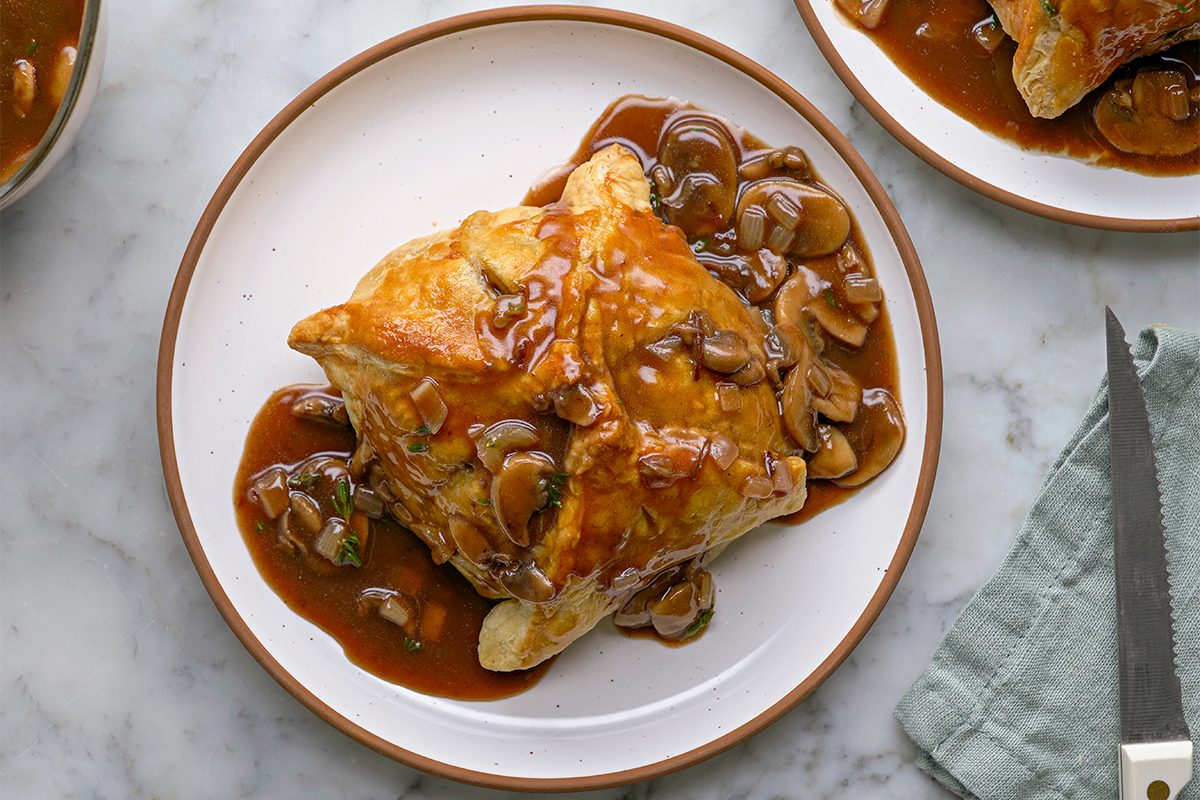 Overhead shot of individual Beef Wellingtons topped with mushroom gravy on a white plate with a brown rim, set on a marble surface beside a green napkin and cutlery;