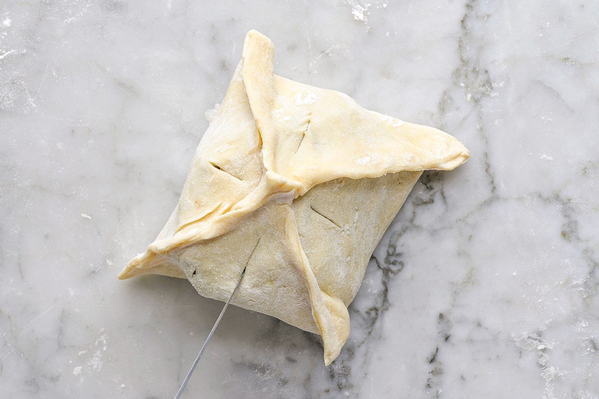 Overhead shot of a hand holding a knife, scoring slits into a square-shaped pastry with folded corners on a marble countertop;