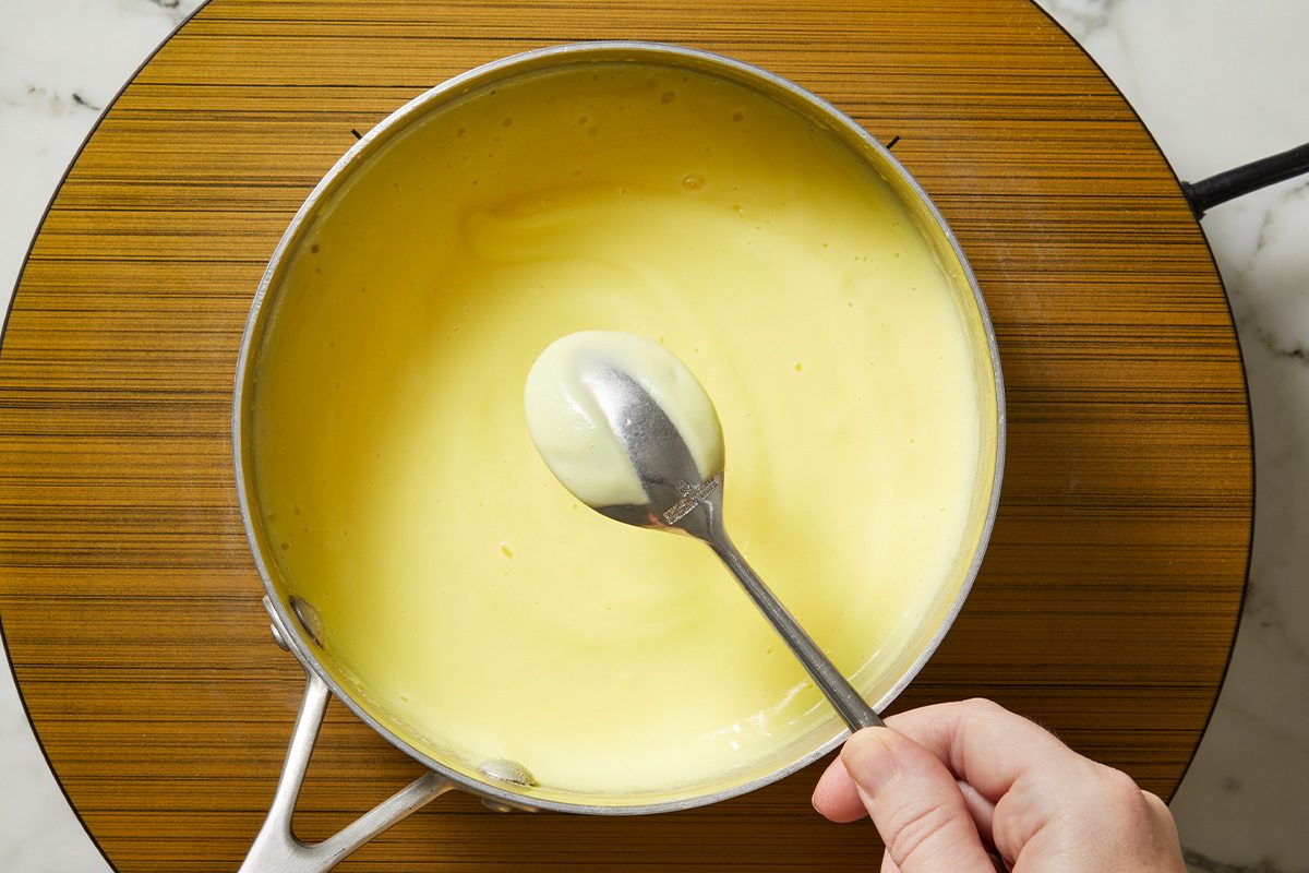 Overhead shot of a hand stirring a smooth, pale yellow sauce or custard with a spoon in a saucepan placed on a round wooden board atop a marble surface;