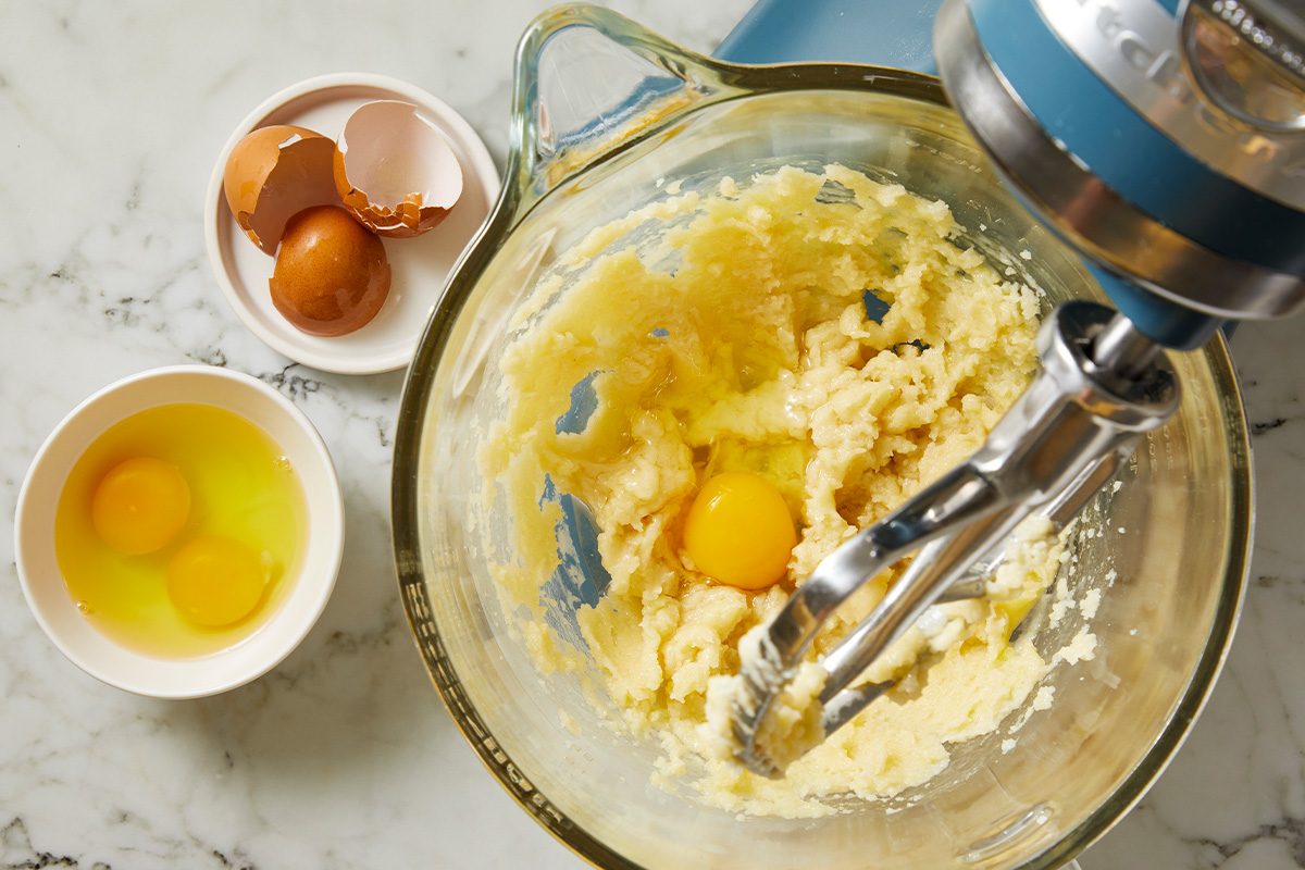 Overhead shot of a stand mixer with a glass bowl containing creamed butter and sugar with one cracked egg; Nearby, a small bowl holds two eggs, and another bowl contains empty eggshells, all on a marble countertop;