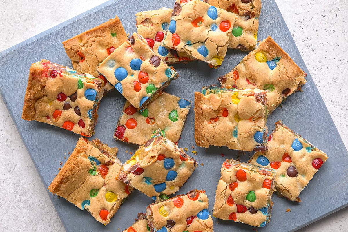 Twelve square cookie bars topped with colorful candy-coated chocolates are arranged on a gray surface. The bars have a golden-brown crust and are scattered in a casual, overlapping pattern.