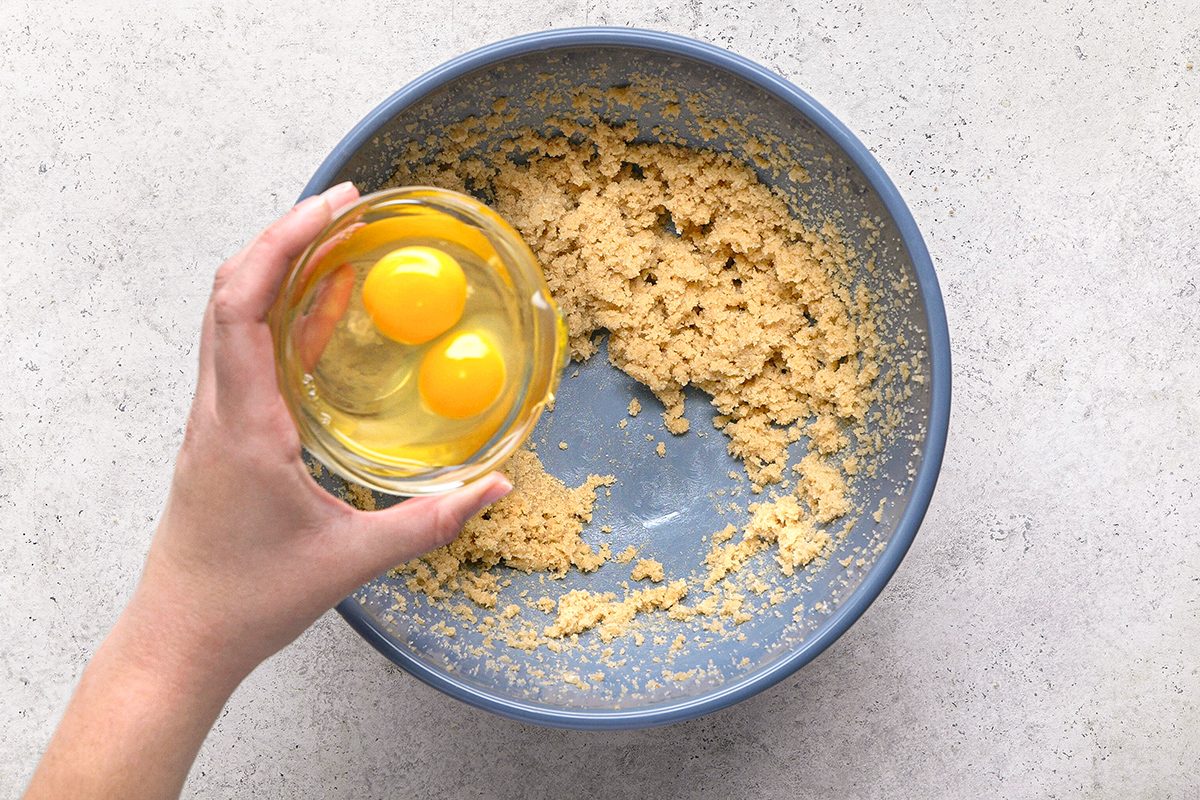 A hand holds a bowl with two cracked eggs above a blue mixing bowl containing partially mixed cookie dough on a light countertop.