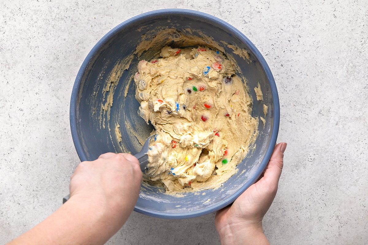 A person is mixing cookie dough with colorful candy pieces in a blue bowl using a spatula, on a light-colored countertop.