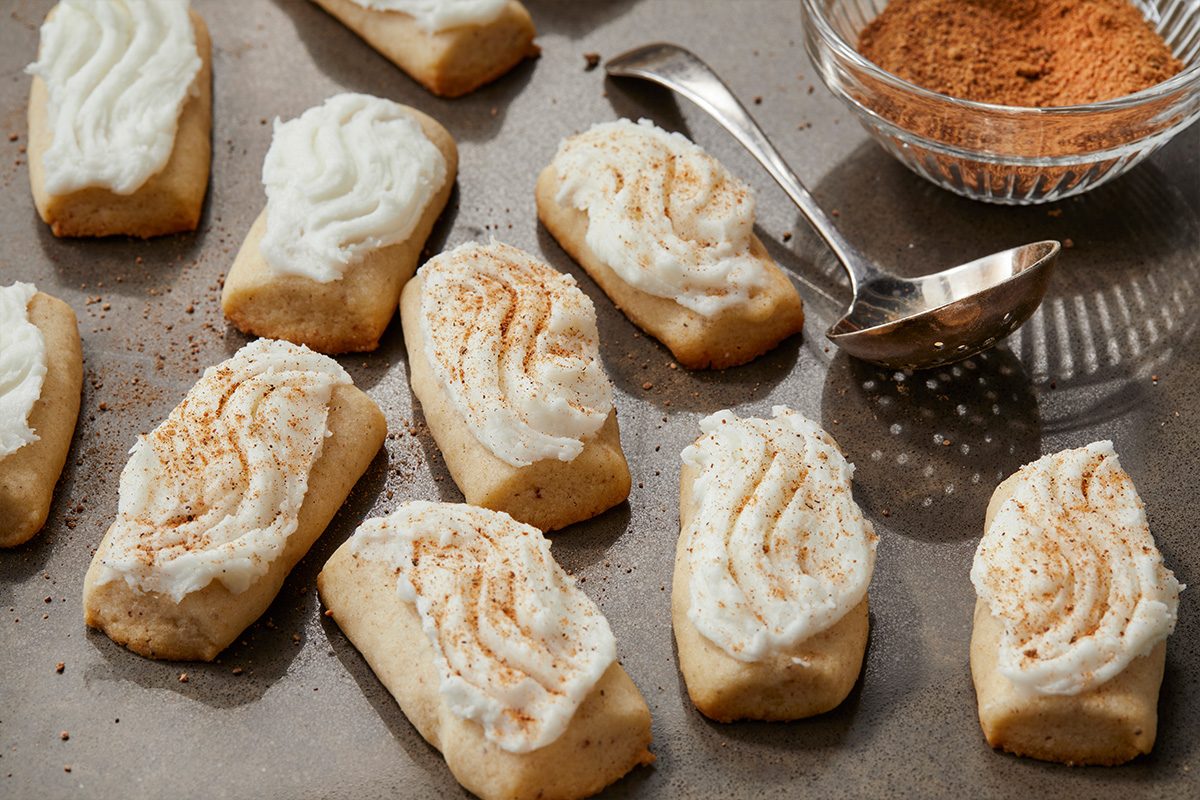 3/4 angle view shot of Frosted Nutmeg Log Cookies, rectangular with piped white frosting and dusted with brown spice, with a spoon and bowl of spice nearby;