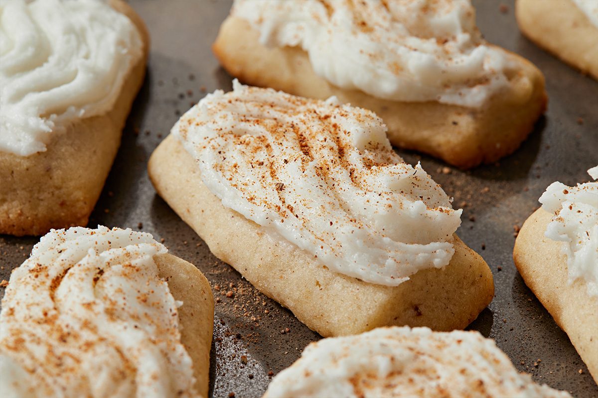Closeup shot of Frosted Nutmeg Log Cookies, rectangular sugar cookies with swirled white frosting and a dusting of cinnamon, set on a baking sheet;