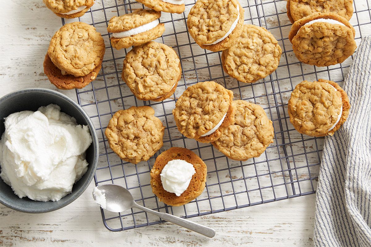 Overhead shot of freshly baked Oatmeal Sandwich Cookies cooling on a wire rack, with a bowl of white cream filling and a kitchen towel on the side.