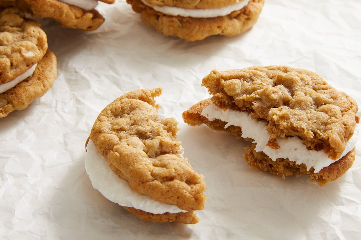 Close-up of assembled Oatmeal Sandwich Cookies with a bite taken out of one, revealing the creamy filling inside. Cookies rest on a crinkled white surface.