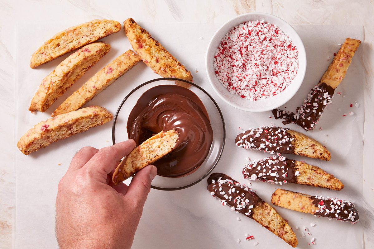 Overhead shot of baked biscotti being dipped into melted chocolate beside bowls of crushed peppermint and finished pieces.