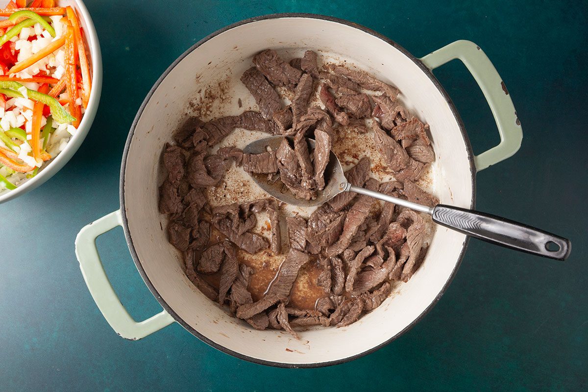 Overhead shot of sliced beef strips browning in a large, light-colored pot with a metal spoon, with a bowl of sliced onions and bell peppers visible to the side on a green surface;