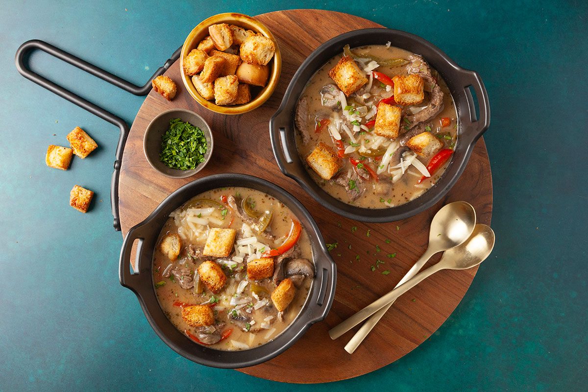 Overhead shot of two bowls of Philly Cheesesteak Soup filled with vegetables, meat, cheese, and croutons, set on a board with extra croutons, fresh herbs, and gold spoons nearby;