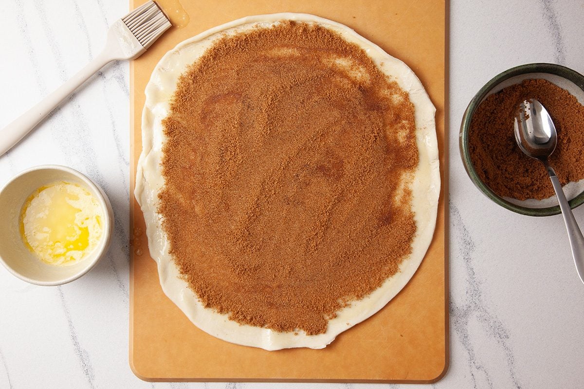 Overhead shot of a round sheet of rolled-out dough on a board coated with a brown cinnamon-sugar mixture; Beside it are a bowl of melted butter with a pastry brush and a bowl of extra cinnamon sugar with a spoon;