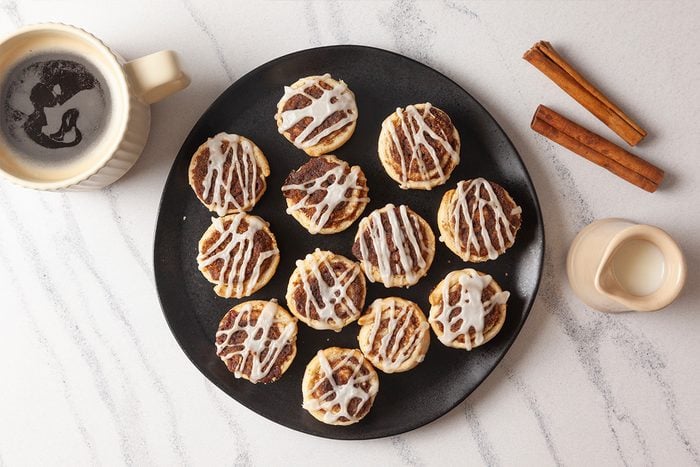 Overhead shot of Pie Crust Cinnamon Rolls drizzled with white icing on a black plate, set beside a cup of coffee with foam art, cinnamon sticks, and a small jug of milk on a marble surface;