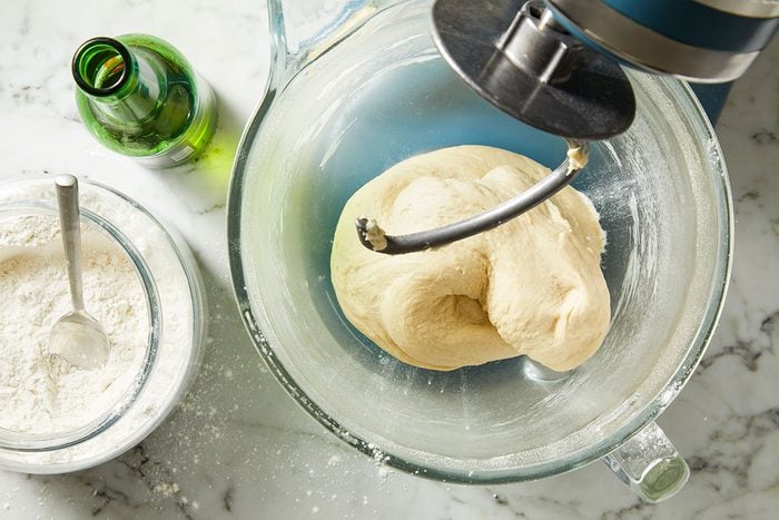 Overhead shot of a glass mixing bowl with dough being kneaded by a stand mixer on a marble countertop, with a bottle and a bowl of flour with a spoon nearby.