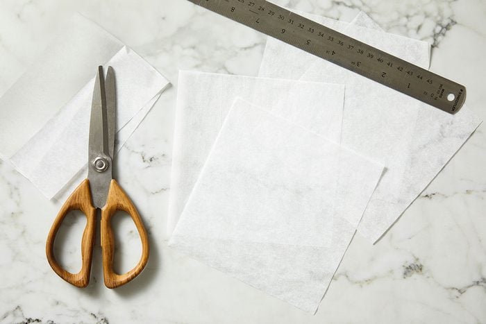 Overhead shot of a pair of scissors with wooden handles, a metal ruler, and several sheets of white wax or parchment paper arranged on a white marble surface.