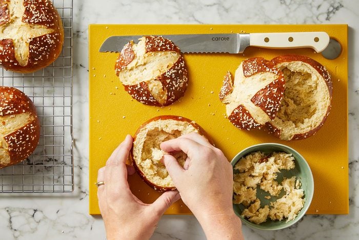 Overhead shot of two hands hollowing out a pretzel bread bowl on a yellow board, with additional bowls, a knife, and a green bowl of crumbs nearby on a marble surface.