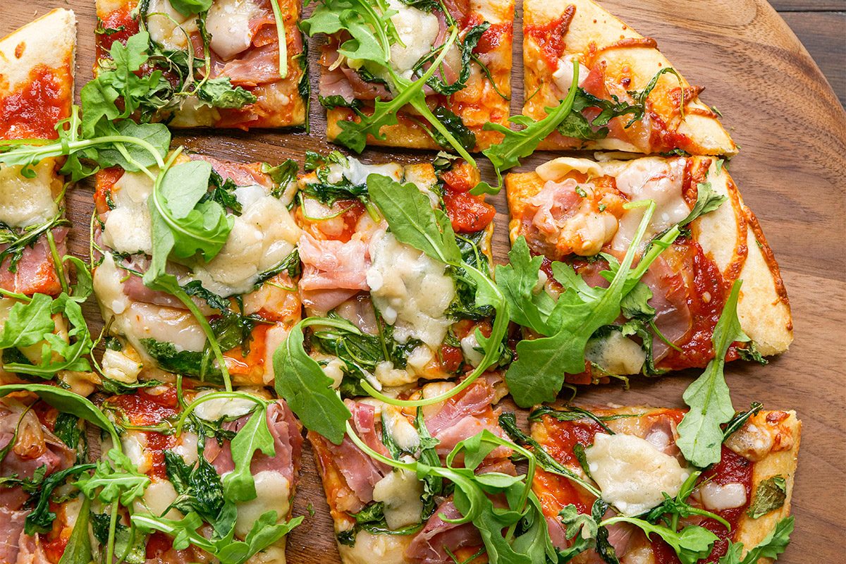 overhead shot of A sliced pizza topped with arugula, melted cheese, tomato sauce, and slices of ham sits on a wooden serving board