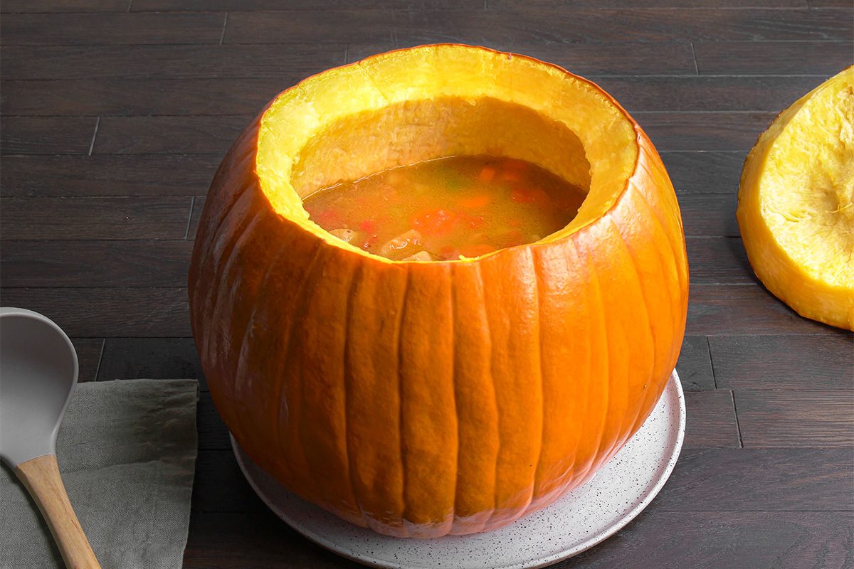 A large, hollowed-out pumpkin filled with soup sits on a plate. The pumpkin's lid is off and resting nearby, and a spoon and napkin are placed beside it on a dark wooden surface.