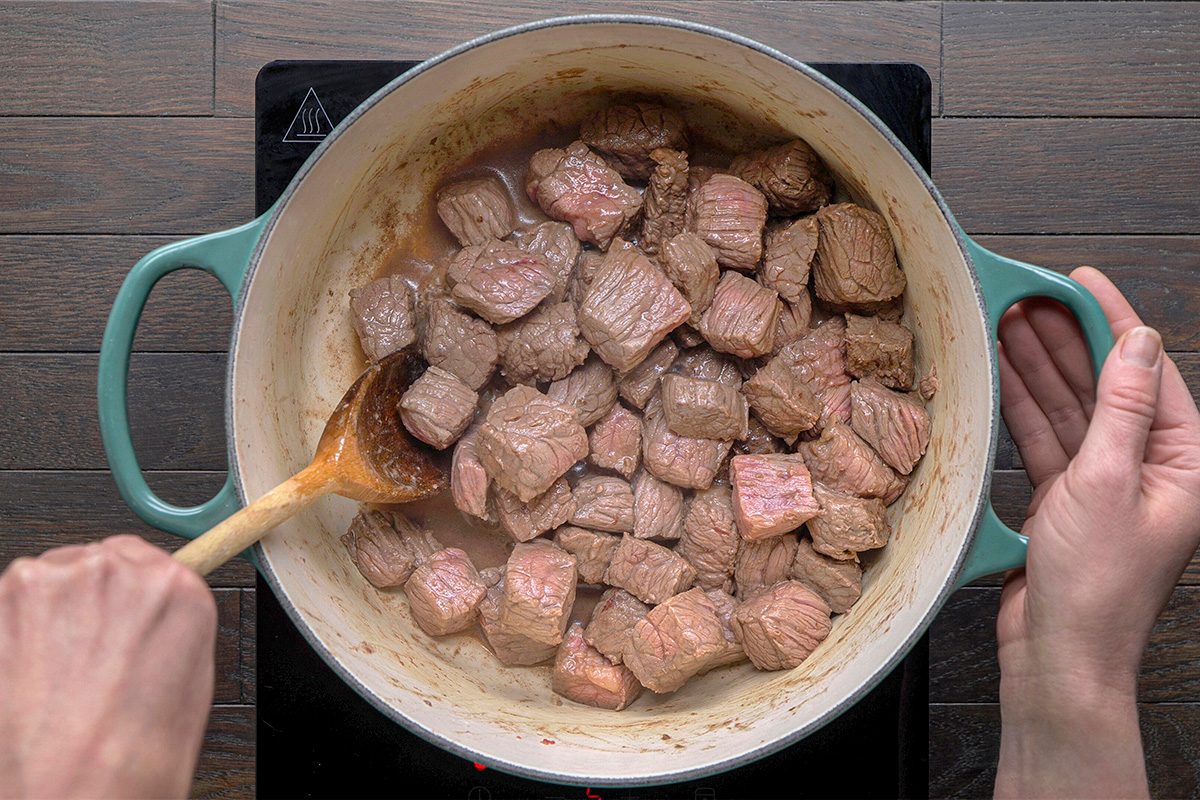 Chunks of beef are being browned in a large green pot on a stovetop, with a person stirring the meat using a wooden spoon.