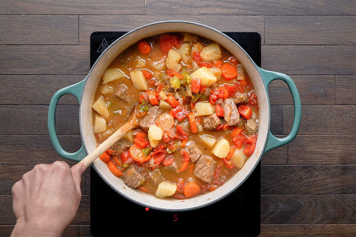 A hand stirs a pot of hearty stew with beef, potatoes, carrots, and other vegetables on a stovetop with a wooden spoon.