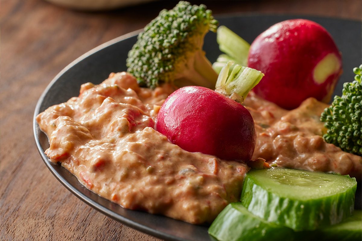 Close-up of Roasted Vegetable Dip served on a dark dish, accompanied by two red radishes, broccoli florets, and cucumber slices;
