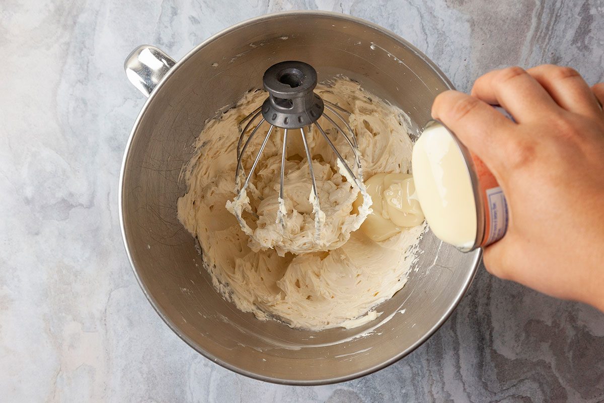 Overhead shot of a hand pouring sweetened condensed milk from a can into a mixing bowl of whipped cream, with a stand mixer fitted with a whisk attachment.