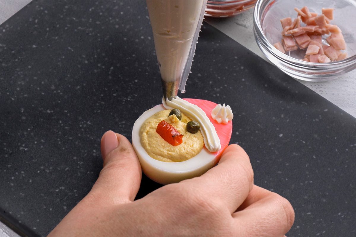 A hand decorates a deviled egg to look like Santa Claus, using a piping bag with white filling for the hat and beard; pieces of red and green toppings and a bowl of chopped ham are nearby.