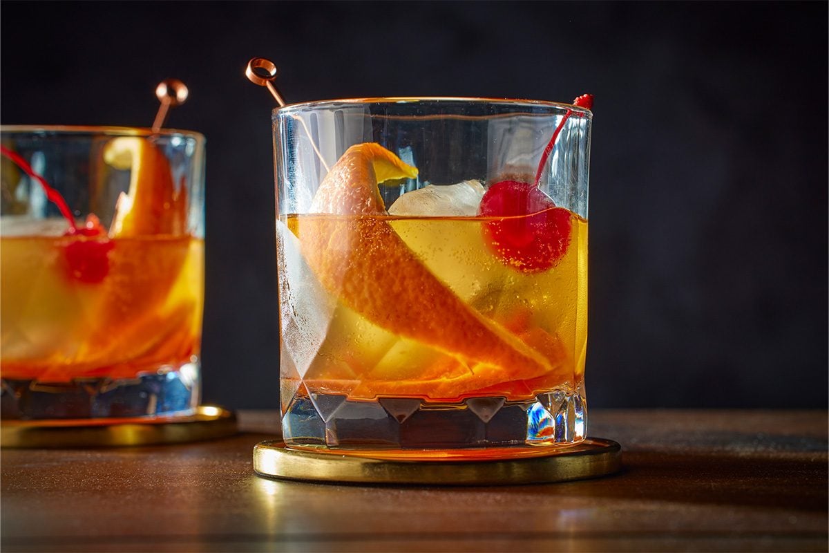 An eye level shot of a Scotch Old-Fashioned, a glass with ice, orange peel, and a maraschino cherry on a cocktail pick, resting on a gold-rimmed coaster, with another drink visible behind;